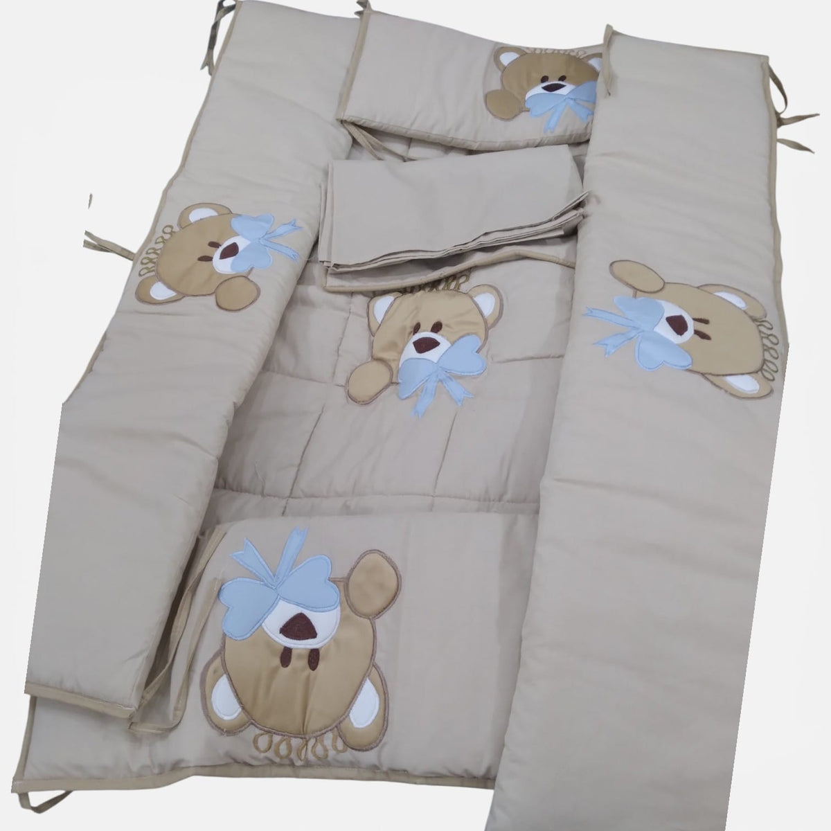 Beige teddy bear cot bedding set laid flat, with embroidered bear and blue bow appliques