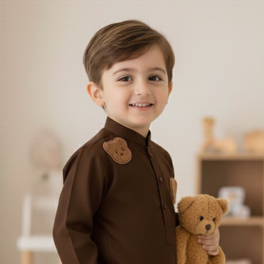 A young boy wearing a dark brown traditional outfit with matching pants and brown shoes, holding a small brown teddy bear in a bright indoor room.